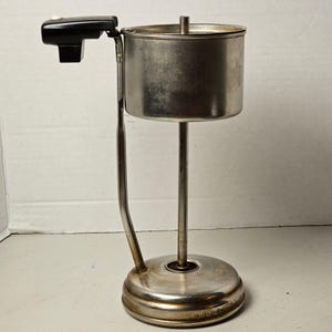 May include: A vintage metal percolator coffee maker component featuring a cylindrical stainless steel basket, a central vertical rod, and a circular base. A black plastic handle with a white button extends from the side of the basket.