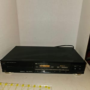 Puede incluir: Un reproductor de discos compactos negro Sharp modelo DX-610 con pantalla digital, botones de reproducción y pausa, y una bandeja para discos. El panel frontal incluye texto como Sharp, Compact Disc Player DX-610, 3-Beam, 20-Track y Digital. Se ve una regla.