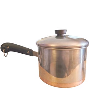 May include: A stainless steel saucepan with a copper-coloured base, a black handle featuring a small metal hanging loop, and a matching stainless steel lid with a black knob. The pot has a cylindrical body and a polished, reflective finish.