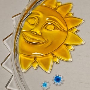 May include: A yellow glass sun with a smiling face, surrounded by clear glass rays. Two small, star-shaped glass ornaments, one blue and one light blue, are at the bottom. The sun is a decorative item.