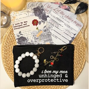 May include: A black canvas pouch with the text "I love my men unhinged & overprotective" and a white beaded bracelet. The pouch has a gold zipper and a keychain with charms. Cards with floral and text designs are in the background.