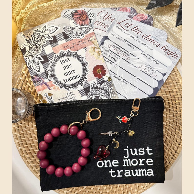 May include: A black canvas pouch with the text "just one more trauma" and a burgundy beaded bracelet. Decorative cards with floral and butterfly designs are also present, along with a keychain.