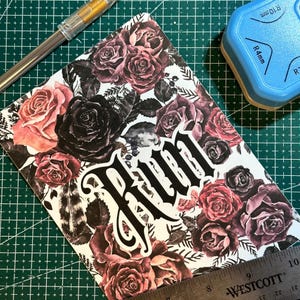 Puede incluir: Una tarjeta blanca con un patr&oacute;n floral con rosas rojas oscuras y negras. La palabra "Run" est&aacute; escrita en letras g&oacute;ticas negras grandes.