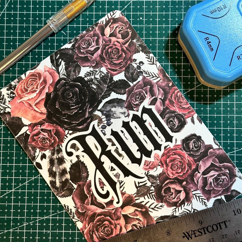 Puede incluir: Una tarjeta blanca con un patr&oacute;n floral con rosas rojas oscuras y negras. La palabra "Run" est&aacute; escrita en letras g&oacute;ticas negras grandes.