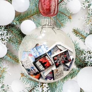 Puede incluir: Un adorno navideño de vidrio transparente lleno de libros de bolsillo en miniatura. Los libros tienen títulos como "Pucking Sweet", "Hideaway", "Cruel Prince", "Effect", "Prom Blood Ash", y "Seven Wings of Night".