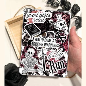 A hand holding a Kindle case insert featuring a dark floral collage design with deep red and black roses. A Dark romance themed reading accessory with "good girls read" and "trigger warnings" text. A bookish gift for dark romance readers.