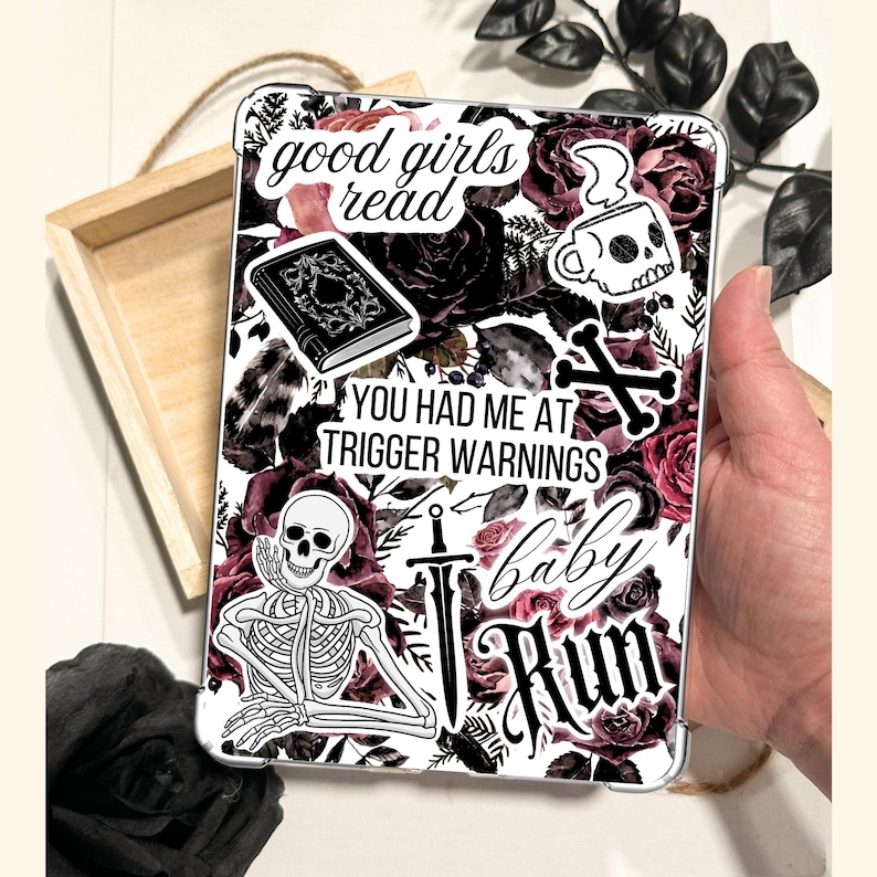 A hand holding a Kindle case insert featuring a dark floral collage design with deep red and black roses. A Dark romance themed reading accessory with "good girls read" and "trigger warnings" text. A bookish gift for dark romance readers.