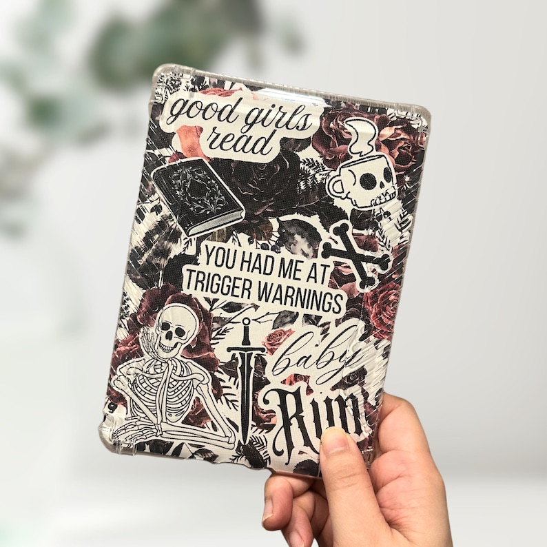May include: A clear protective case for an e-reader, decorated with stickers. The stickers include a skull, roses, a book, and the phrases "good girls read" and "You had me at trigger warnings".