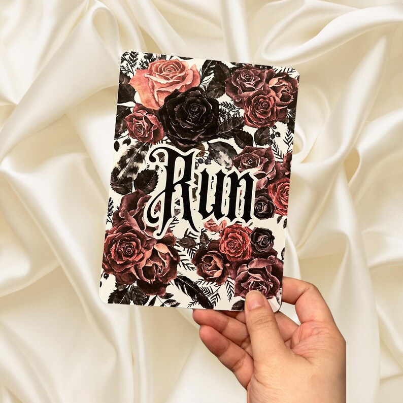 Puede incluir: Una tarjeta blanca con un dise&ntilde;o floral con rosas de color rojo oscuro, rosa y negro. La palabra "Rum" est&aacute; escrita en letras g&oacute;ticas negras.