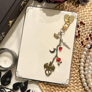 May include: A clear, rectangular case with a gold heart-shaped clasp and a charm featuring a crown, a crescent moon, a sword, a dragon, and a red rose. The case is on a white surface.