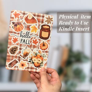 May include: A decorative Kindle insert featuring fall-themed illustrations. The design includes pumpkins, maple leaves, a slice of pie, and the words "hello FALL". The insert is held by a hand and has a plaid background.