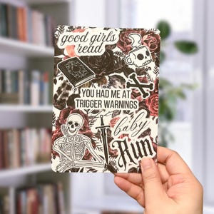 May include: A white card with a floral background and various black and white stickers. The stickers include a skull, a sword, a skeleton, a book, and text that reads "good girls read", "you had me at trigger warnings", and "baby run".