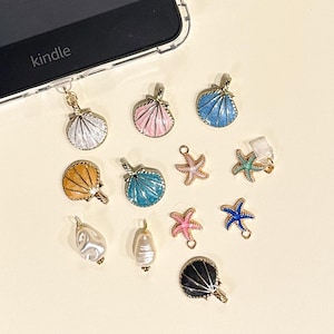 May include: A collection of seashell and starfish charms in various colors, including white, pink, blue, and gold. Also included are two pearl-shaped charms and a black fan-shaped charm. The word "kindle" is visible on a black electronic device.