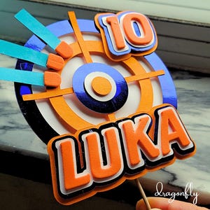 May include: A cake topper featuring a blue and orange target with a white bullseye, a blue and orange number 10, and the name Luka in orange letters with black outlines.