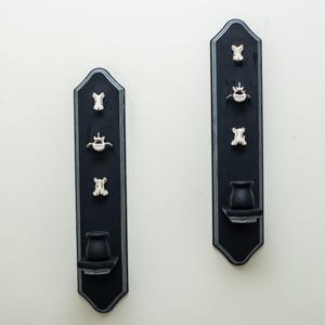May include: Two black wall-mounted candle holders with silver-painted edges. Each vertical plaque features three small, white, bone-like vertebrae ornaments attached above a circular candle cup base, creating a symmetrical display of dark decor.