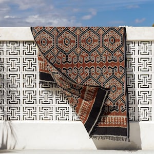 May include: A rectangular textile featuring a repeating geometric pattern in rust orange, black and cream. The fabric displays a symmetrical hexagonal motif with intricate linework and a solid black border along the edges, finished with short fringe.
