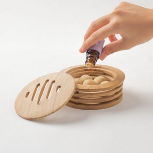 Puede incluir: Un difusor de aromaterapia de madera con un diseño apilable. El difusor tiene una tapa con ranuras recortadas y contiene varias bolas de madera pequeñas y redondas. Una mano vierte aceite esencial de una pequeña botella marrón en el difusor.