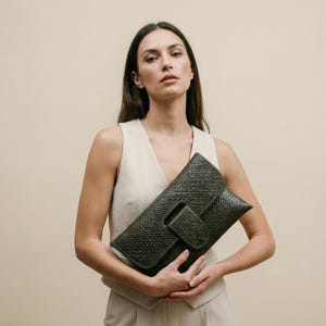 May include: A dark olive green woven clutch bag with a rectangular flap closure. The bag features a textured, basket-weave pattern throughout the exterior surface, providing a structured appearance with a secure front flap and a matching woven tab.