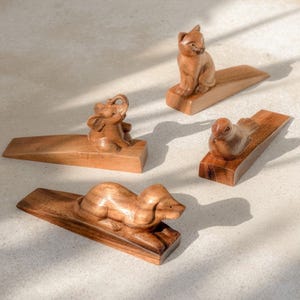 May include: Four wedge-shaped door stops carved from light brown wood, each featuring a different animal figure on top: a sitting cat, a small elephant with its trunk raised, a bird, and a dog lying down in a resting position.