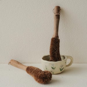 May include: Two wooden handled dish brushes with brown bristles. One brush is resting on a white surface, while the other is inside a cream-colored mug decorated with floral designs. The brushes have a natural, eco-friendly aesthetic.
