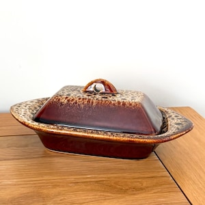 May include: A rectangular ceramic butter dish featuring a deep brown base colour with a speckled cream and tan glaze pattern on the lid and the wide, flat rim. The lid includes a small, arched handle centred on the top for easy lifting.