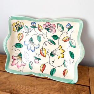 May include: A rectangular ceramic serving tray with a cream base and a light green border. The tray is decorated with colourful floral and leaf designs in pink, yellow, blue, and green. The tray is placed on a wooden surface.
