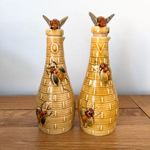 May include: Two ceramic bottles with bee motifs. The bottles are light brown with a textured, honeycomb pattern. Each has a bee-shaped stopper and bee illustrations. The bottles are displayed on a wooden surface.