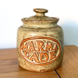 May include: A ceramic marmalade jar with a lid. The jar is beige with a brown oval that reads "MARMALADE" in raised orange letters. The jar is sitting on a wooden surface.