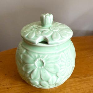May include: A pale green ceramic jar with a lid, decorated with embossed daisy patterns. The lid has a small handle and a spoon notch. The jar sits on a wooden surface. The jar is approximately 10 cm in height.
