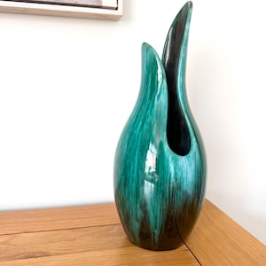 May include: A tall, teardrop-shaped ceramic vase featuring a glossy finish with a blend of deep emerald green and black colours. The vase has a smooth, curved silhouette with an asymmetrical opening at the top, showcasing a dark interior.