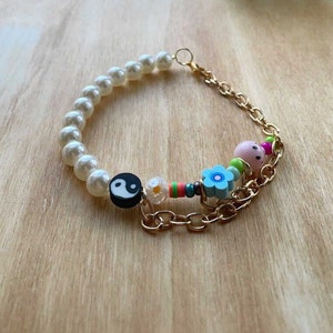 May include: A gold chain bracelet with white pearl beads, a yin yang charm, a flower charm, and a smiley face charm.
