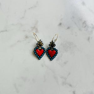 May include: A pair of heart-shaped earrings with a red center, outlined with turquoise beads. The earrings have a gold-colored top with a flame design and gold-colored hoop closures. The earrings are displayed on a white marble surface.
