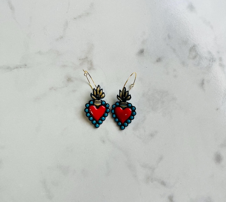May include: A pair of heart-shaped earrings with a red center, outlined with turquoise beads. The earrings have a gold-colored top with a flame design and gold-colored hoop closures. The earrings are displayed on a white marble surface.