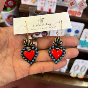 May include: A pair of handmade earrings featuring a red heart design with turquoise accents and gold-colored accents. The earrings are part of the "birdie & clay" handmade accessories collection.