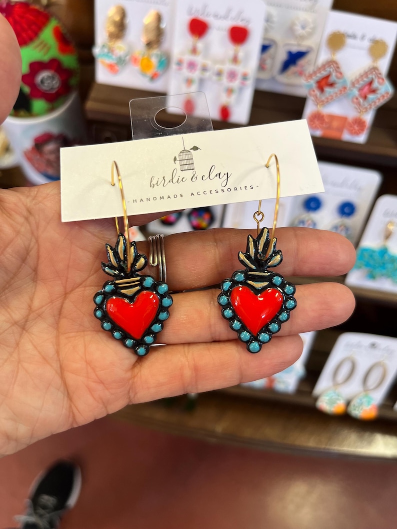 May include: A pair of handmade earrings featuring a red heart design with turquoise accents and gold-colored accents. The earrings are part of the "birdie & clay" handmade accessories collection.