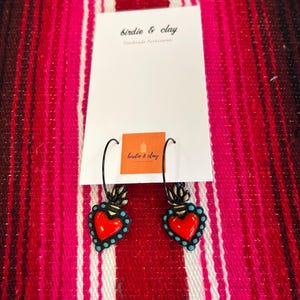 May include: A pair of heart-shaped earrings with a red center and turquoise accents. The earrings are black and have a decorative design at the top. The earrings are displayed on a white card with the text "birdie & clay Handmade Accessories".