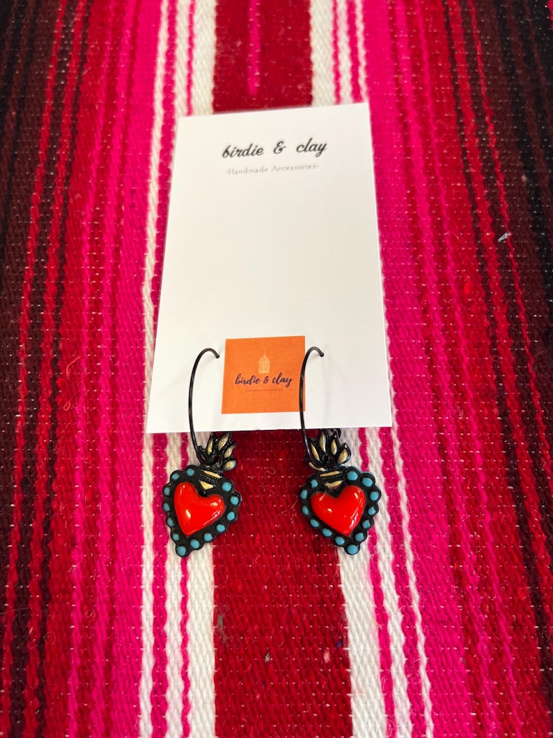 May include: A pair of heart-shaped earrings with a red center and turquoise accents. The earrings are black and have a decorative design at the top. The earrings are displayed on a white card with the text "birdie & clay Handmade Accessories".