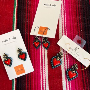 May include: Earrings with a red heart design, turquoise accents, and gold-colored flames. The earrings are displayed on white cards with the text "birdie & clay Handmade Accessories". The background is a red and pink striped textile.