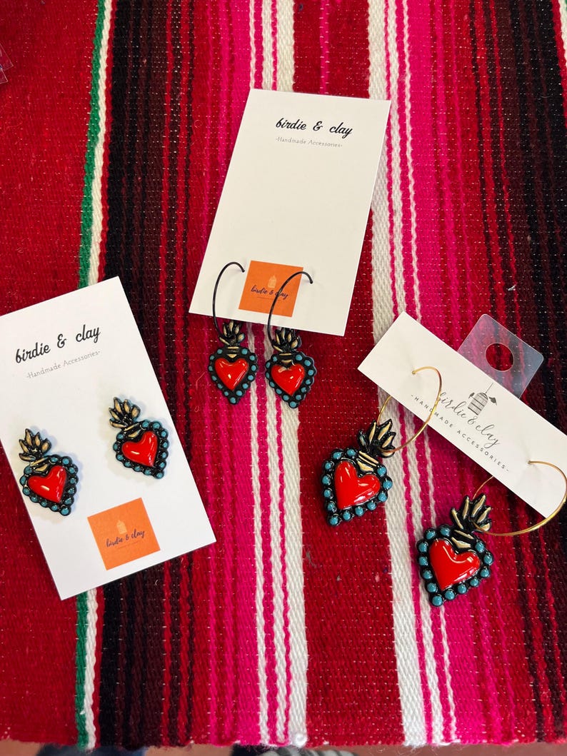 May include: Earrings with a red heart design, turquoise accents, and gold-colored flames. The earrings are displayed on white cards with the text "birdie & clay Handmade Accessories". The background is a red and pink striped textile.