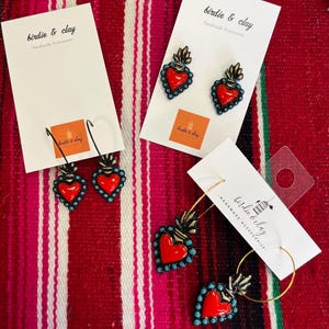 May include: Earrings with a red heart design, outlined in turquoise beads, and a gold flame detail. The earrings are displayed on white cards with the text "birdie & clay Handmade Accessories". The background is a red and white striped textile.