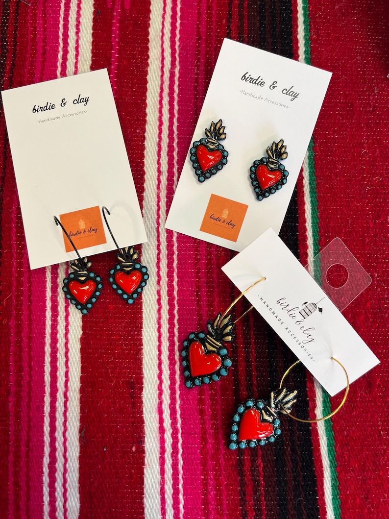 May include: Earrings with a red heart design, outlined in turquoise beads, and a gold flame detail. The earrings are displayed on white cards with the text "birdie & clay Handmade Accessories". The background is a red and white striped textile.