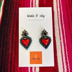 May include: A pair of handmade heart-shaped earrings with a red center, teal border, and gold accents. The earrings are displayed on a white card with the text "birdie & clay - Handmade Accessories" and a logo. The background is a red and white striped textile.