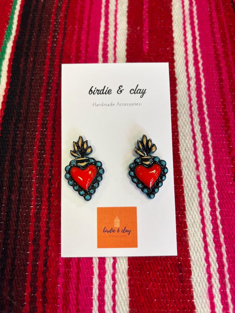 May include: A pair of handmade heart-shaped earrings with a red center, teal border, and gold accents. The earrings are displayed on a white card with the text "birdie & clay - Handmade Accessories" and a logo. The background is a red and white striped textile.