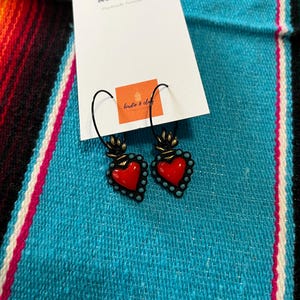 May include: A pair of heart-shaped earrings with a red center and turquoise bead border. The earrings are suspended from black hooks and are displayed on a white card with the text "birdie & clay" and a small orange square with the same text. The background is a turquoise and multicolored woven textile.
