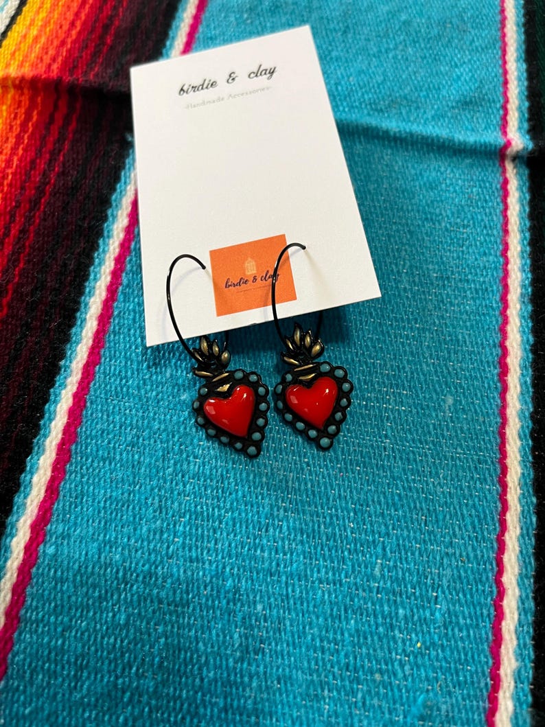 May include: A pair of heart-shaped earrings with a red center and turquoise bead border. The earrings are suspended from black hooks and are displayed on a white card with the text "birdie & clay" and a small orange square with the same text. The background is a turquoise and multicolored woven textile.