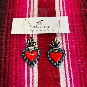 May include: A pair of gold hoop earrings with red heart pendants. The heart pendants are outlined in black with turquoise bead accents. The earrings are handmade accessories. The text on the tag reads "birdie & clay - HANDMADE ACCESSORIES".