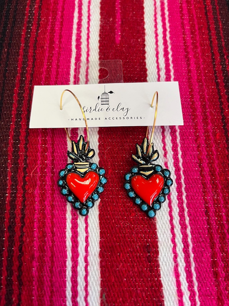 May include: A pair of gold hoop earrings with red heart pendants. The heart pendants are outlined in black with turquoise bead accents. The earrings are handmade accessories. The text on the tag reads "birdie & clay - HANDMADE ACCESSORIES".