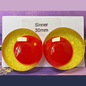 May include: A pair of circular earrings featuring a bright yellow glitter base with a smooth, solid red circular accent layered on top. The white display card behind the earrings is printed with the text Sinner 30mm, indicating the size.