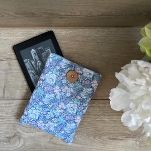 May include: A rectangular fabric sleeve featuring a dense floral pattern in shades of blue, purple, and white, secured with a single round wooden button and an elastic loop, partially covering a black e-reader device with a screen display.