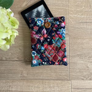 May include: A navy blue quilted e-reader case with a floral and butterfly design. It has a wooden button closure and a colourful pattern with red, blue, and purple flowers. The e-reader is partially visible.